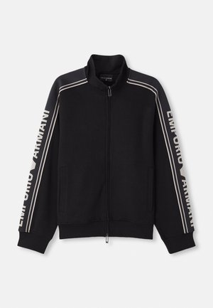 Veste noire zippée avec col haut, présentant des rayures beiges et le texte "Emporio Armani" le long des manches. Tissu doux, deux poches latérales.