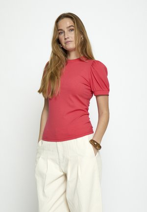 Jeune femme aux longs cheveux portant un haut rouge à manches bouffantes, un pantalon large crème et un bracelet en bois, posant les mains dans les poches.