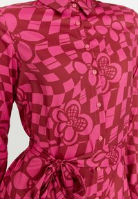 Červená košile s růžovým geometrickým a květinovým vzorem, s límečkem, předním zapínáním na knoflíky a zdůrazněním v pase pomocí zavázání.