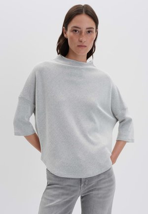 Femme aux cheveux bruns raides portant un haut en tricot gris clair ample à manches mi-longues et un jean gris clair, se tenant devant un fond uni.