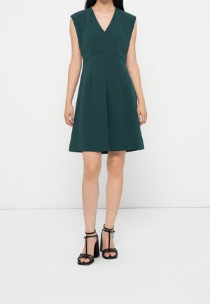 Woman wearing sleeveless dark green knee-length dress with V-neck and black high-heeled strappy sandals, standing against plain white background.