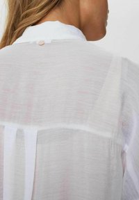 Sheer white blouse with a collared neckline, visible stitching, and a button closure at the back. Features delicate, textured fabric.