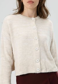 Beige knitted cardigan with a ribbed hem, featuring five white buttons down the front. Soft texture, cropped design.
