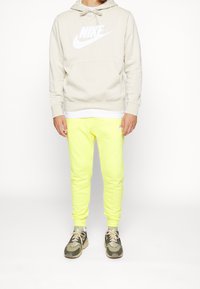 Béžová mikina Nike s přední kapsou a bílým logem, doplněná jasně žlutými tepláky a hnědými sportovními botami s černými detaily.