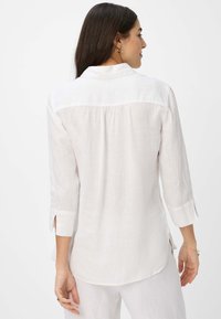 White linen shirt with long sleeves, a back pleat, and side slits, featuring a collar and a relaxed fit. Soft texture and lightweight design.