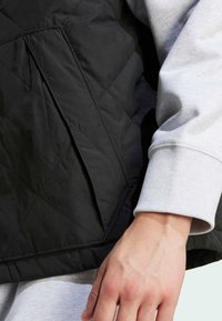 Crni stegnuti prsluk s geometrijskim uzorkom i kutnim džepom, složen preko svijetlosivog majice dugih rukava s rebrastim manžetama.