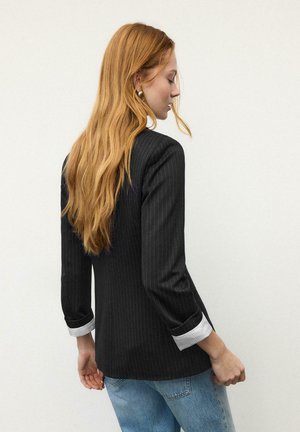 Blazer noir à fines rayures avec une coupe ajustée et des manches trois-quarts, doté de poignets blancs. Le modèle porte un jean bleu clair.