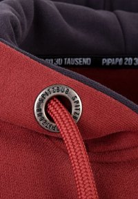 Rood en donkergrijs stof met een gestructureerd oppervlak. Bevat een metalen oogje met "SPITZBUB" gegraveerd en een geweven rode trekkoord.