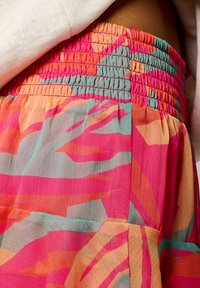 Jupe colorée et légère avec une taille froncée. Le tissu présente un motif floral dans des teintes de rose, orange et turquoise. Texture douce et transparente.