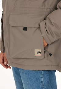 Beige jakke lavet af struktureret stof, med en stor frontlomme, et logo-patch og en sort lynlås i kanten. Bæres med blå jeans.