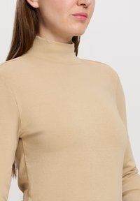 Beżowa bluzka typu golf wykonana z miękkiego, elastycznego materiału. Posiada wysoki kołnierz oraz dopasowany krój, z bezszwowymi rękawami do łokci.