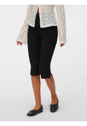 Femme portant un pantalon noir court, un haut blanc en dentelle à manches longues et des ballerines noires, debout devant un fond uni.