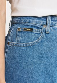 Vaaleansiniset farkut klassisella istuvuudella. Etutaskussa näkyy kultakirjailtu logo ja edessä on metalliset niitit.