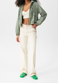 Groene lichtejack, witte cropped top, beige wijde broek en groene gesloten schoenen. De stoffen lijken glad; het ontwerp is casual.
