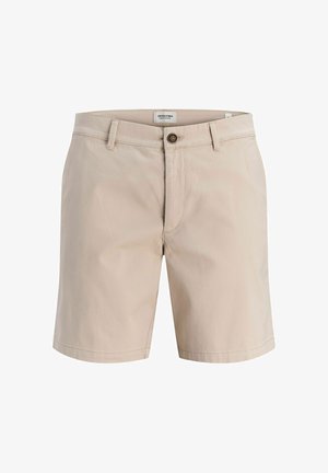Gutt står med hendene i lommer, iført en svart kortermet polo, beige shorts, kremfargede sokker og beige sko på en ensfarget bakgrunn.