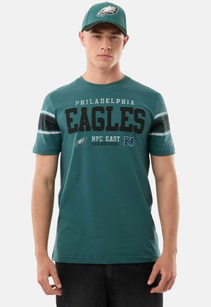 Giovane uomo che indossa un cappellino verde dei Philadelphia Eagles e una maglietta abbinata con il nome della squadra e i loghi, in piedi davanti a uno sfondo semplice.