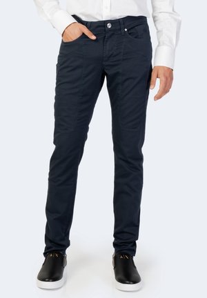 Mann trägt dunkelblaue, schmal geschnittene Hose mit diagonalen Vordertaschen und schwarze Slip-on-Schuhe mit weißen Sohlen.