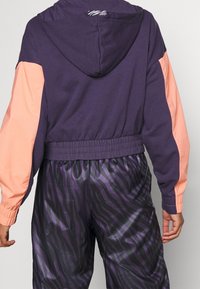 Fialová krátká mikina s korálovými rukávy; má kapuci a elastický pas. Kombinována s černými kalhotami se zebřím vzorem.