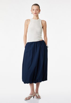 Kvinde står med hænderne i lommerne iført hvid ærmeløs ribbet top, navy midi nederdel, hvide sandaler med hæl og sølvfarvede øreringe i hulring.