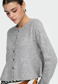 Sivi kardigan od mekog, teksturiranog materijala; ima okrugli izrez, duge rukave i gumbe sprijeda. Značajan detalj prošivanja na rubu.