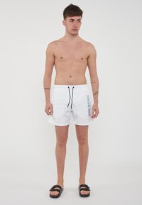 Witte zwembroek met een trekkoord in de taille, met een blauw logo-patroon aan de zijkant. Het model is blote voeten en draagt zwarte sandalen.