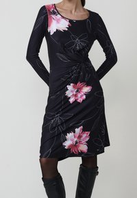 Femme portant une robe noire mi-longue avec imprimés floraux roses, manches longues, détail nœud sur le côté, associée à des collants noirs et des bottes montantes.