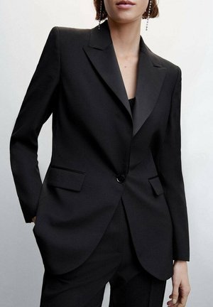 Personne portant un costume noir sur mesure avec un blazer à un bouton, des revers aiguisés et un pantalon noir assorti sur un fond clair uni.