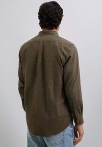 Camisa de cuadros marrón con botones, de manga larga y cuello clásico, hecha de una tela suave. Jeans desgastados de color azul claro visibles abajo.