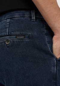 Džínové kalhoty v tmavě modré látce s jemnou strukturou. Obsahují zadní kapsu s etiketou Trussardi a kontrastní švy. Viditelný detail knoflíku.