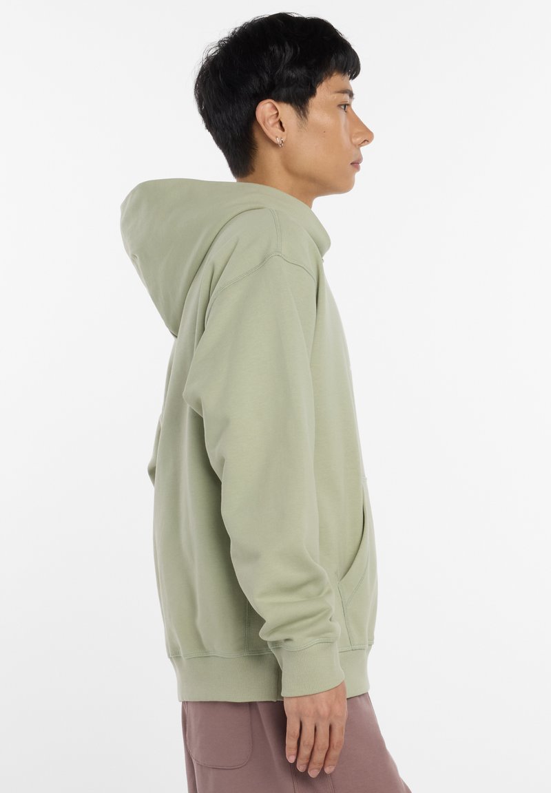 Sweat à capuche vert clair avec cordon de serrage, poche avant et poignets côtelés. Texture douce et coupe décontractée, associé à un pantalon marron clair.