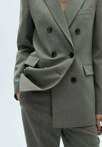 Blazer grigio doppiopetto con grandi bottoni neri, revers strutturati e taschino sul petto; abbinato a pantaloni su misura coordinati.