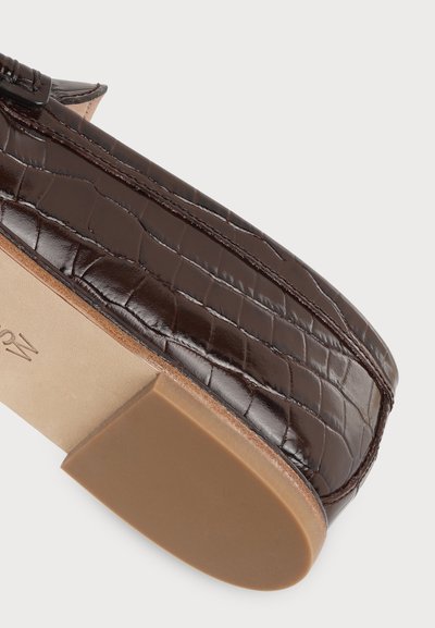 Gros plan d'un mocassin marron à texture motif crocodile et semelle plate beige sur fond blanc.