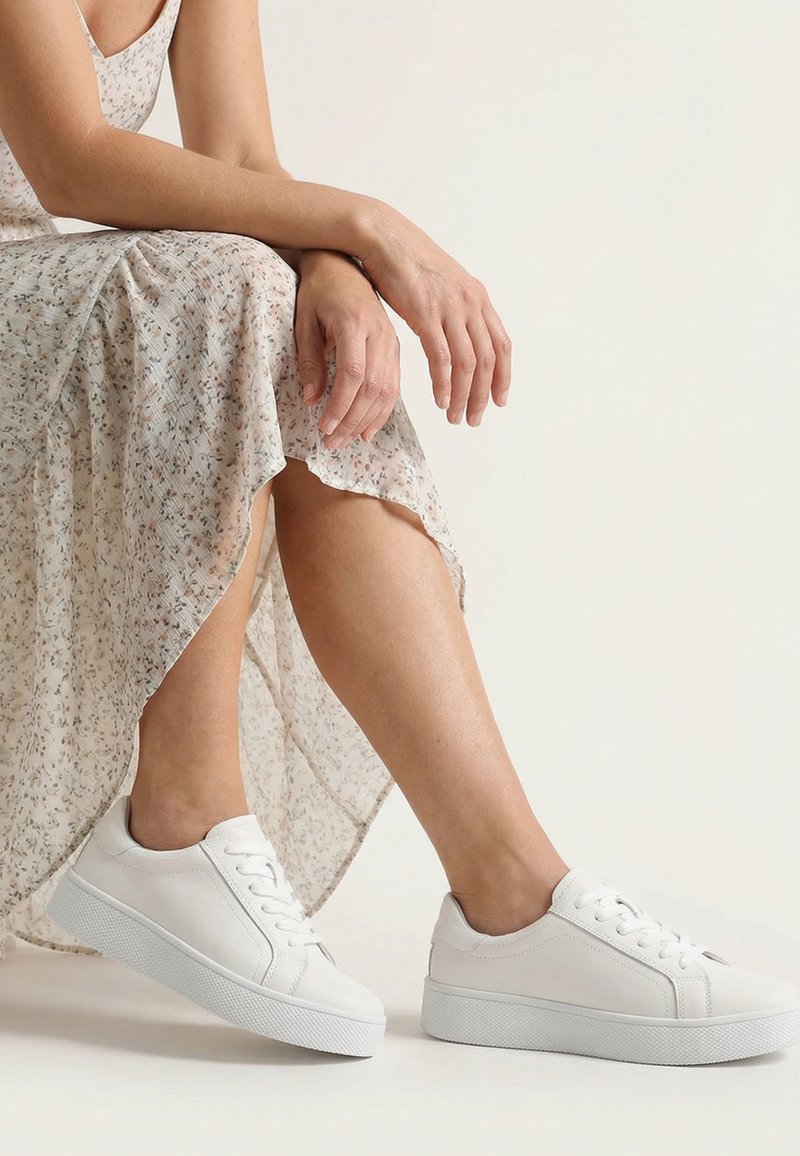 Frau sitzt mit den Händen im Schoß, trägt ein leichtes Blumenkleid und weiße niedrige Sneaker vor einem schlichten Hintergrund.