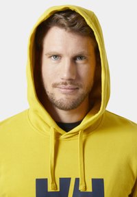 Sudadera amarilla con bolsillo canguro en el frente, capucha ajustable con cordón y un gran logo azul oscuro en el pecho. Textura suave y corte relajado.