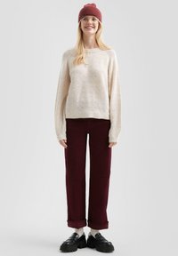 Crème trui met een gestructureerd bladmuster, gecombineerd met bordeauxkleurige corduroy broek. Accessoires zijn een bordeauxkleurige beanie en zwarte chunky loafers.