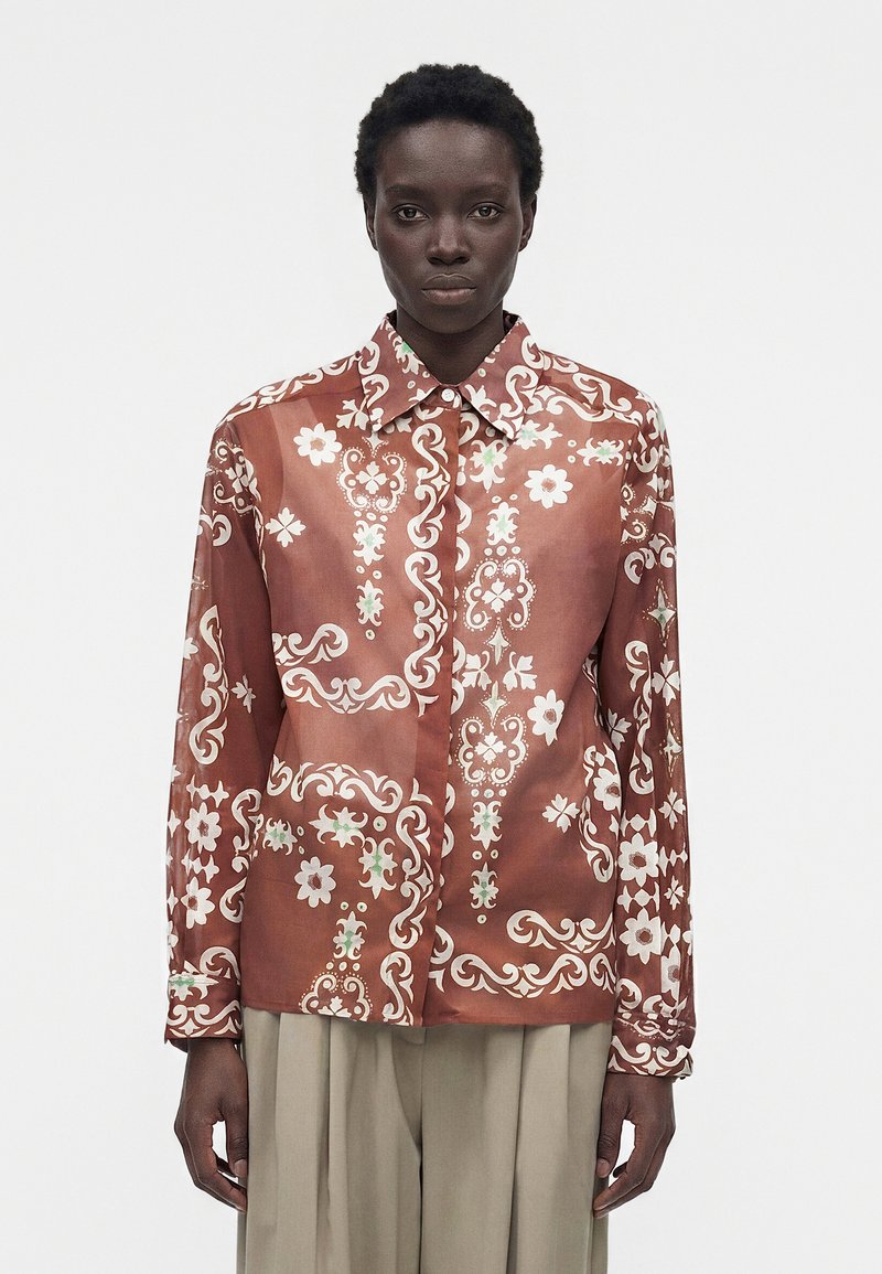 Άτομο με σκουρόχρωμο δέρμα φοράει ένα σκουλαρισμένο, μακρυμάνικο πουκάμισο σε σκουροκόκκινο-πορτοκαλί χρώμα με λευκά λουλουδάτα και στροβιλιστικά μοτίβα, συνδυασμένο με μπεζ πλισέ παντελόνια.