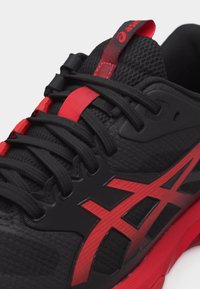 Αθλητικό παπούτσι σε μαύρο και κόκκινο με επάνω μέρος από δίχτυ, μαύρα κορδόνια και κόκκινα σχεδιαστικά στοιχεία, συμπεριλαμβανομένου του λογότυπου στη πλευρά και της ταινίας τράβηγμα στη γλώσσα.