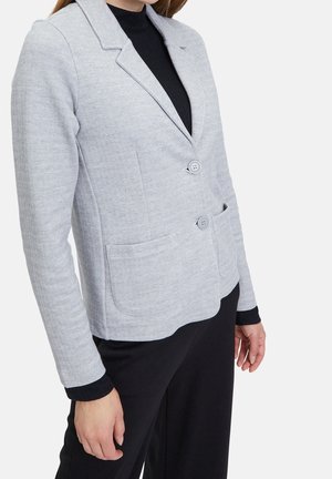 Kobieta ubrana w jasnoszary żakiet z dwoma guzikami i przednimi kieszeniami na czarnej bluzce z długim rękawem i czarnych spodniach, na białym tle.