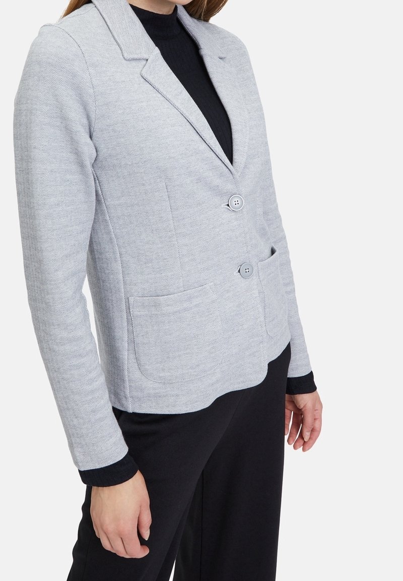 Femme portant un blazer gris clair à deux boutons avec des poches avant sur un haut noir à manches longues et un pantalon noir, sur fond blanc.