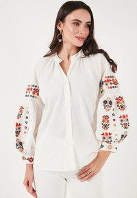 Witte blouse met opgestoken mouwen, voorzien van kleurrijke bloemachtige borduursels op de manchetten. Gemaakt van lichtgewicht stof, getextureerd met verfijnde patronen.