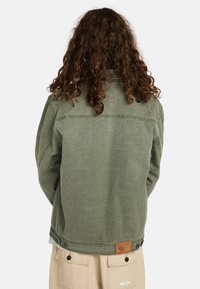 Chaqueta de mezclilla verde oliva con un ajuste relajado, costuras visibles, un parche de cuero en la parte posterior y puños abotonados.