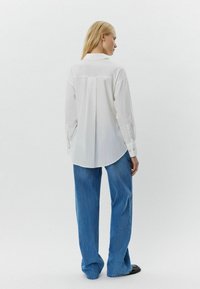 Camicia bianca a maniche lunghe con piega sul retro, orlo curvo e dettagli con bottoni, abbinata a jeans in denim blu larghi, su uno sfondo semplice.