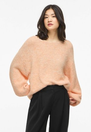 Jeune femme aux cheveux bruns mi-longs portant un pull ample de couleur pêche et un pantalon noir taille haute, sur un fond uni.