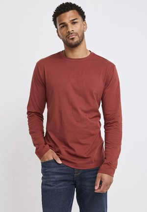 Langarm-T-Shirt in Rostfarbe aus weichem Baumwollstoff, mit Rundhalsausschnitt und schlanker Passform, kombiniert mit dunkelblauen Jeans.