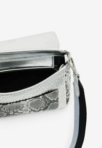 Srebrna torebka o fakturze imitującej wężową skórę, z zamkiem błyskawicznym u góry i czarnym paskiem. Wewnątrz wykończona gładką, srebrną podszewką.