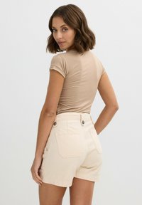 Top ajustado beige de manga corta combinado con pantalones cortos beige claro. Los pantalones cortos cuentan con dos bolsillos traseros y acentos de botón. Textura suave.
