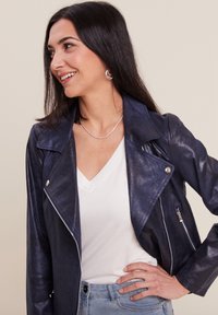 Giacca biker in ecopelle blu navy con accessori in argento, zip asimmetrica e colletto rialzato, indossata sopra una maglietta bianca a scollo a V e jeans chiari.