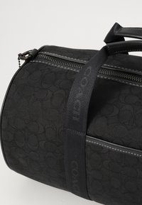 Schwarze Reisetasche mit eingeprägtem Logomuster, ausgestattet mit einem Reißverschluss an der Oberseite, strukturiertem Oberflächenmaterial und Stoffgriffen mit einem glatten Akzentriemen.