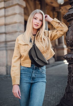 Černá kožená crossbody taška s zakřiveným designem a výrazným logem, nošená přes béžovou semišovou bundu a světle modré džíny. Hladký povrch.