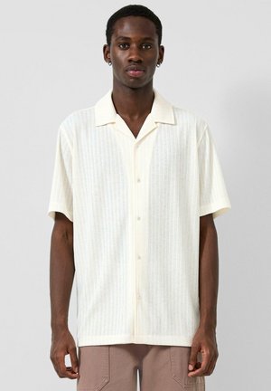 Giovane uomo con indosso una camicia cremisi a maniche corte con sottili righe verticali, in piedi davanti a uno sfondo chiaro e semplice.